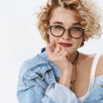 Giovane donna bionda riccia con occhiali e giacca di jeans sorride poggiando ...