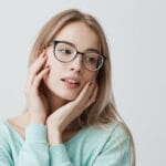 Giovane donna bionda con occhiali e maglione azzurro guarda serena di lato, m...